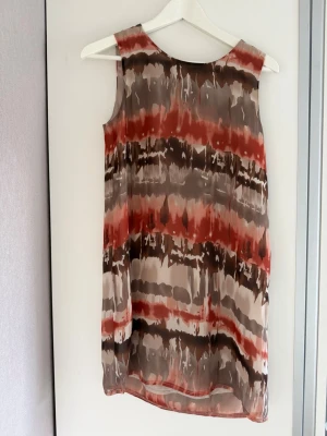 Ärmlös mönstrad klänning i brunt och rött dry lake  - Säljer en ärmlös kortklänning med abstrakt mönster i bruna, röda och beige toner. Klänningen har en rak passform och är tillverkad i ett lätt, luftigt material som passar perfekt för varmare dagar.