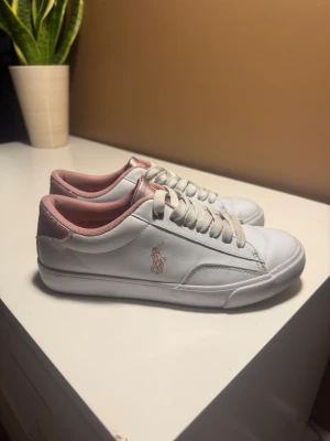 Vita och rosa sneakers från Polo Ralph Lauren - Snygga vita sneakers från Polo Ralph Lauren med rosa detaljer och logga på sidan. Skorna har låga skaft, vita snören och rosa insida samt hälparti. Tillverkade i slätt syntetmaterial med klassisk design.