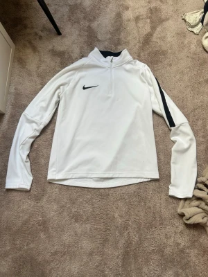 Vit Nike Dri-Fit träningströja - Säljer en vit långärmad träningströja från Nike med svart logga och svart detalj på ena ärmen. Tröjan har halv dragkedja och är tillverkad i Dri-Fit material som andas och transporterar bort fukt. Perfekt för träning eller uppvärmning.