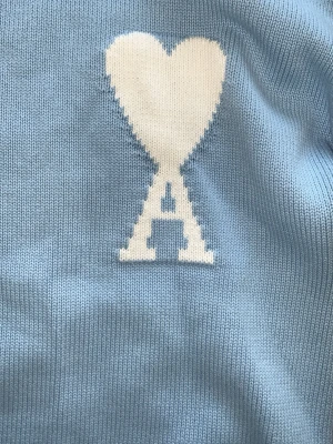 Ljusblå stickad tröja Ami Paris - Ljusblå stickad tröja från Ami Paris med deras ikoniska vita hjärta och A-logga på bröstet. Tröjan har korta ärmar, ribbstickad nederkant och halsringning. Perfekt för en avslappnad men trendig look. OBS! på tröjan står det size L men det är lite större än M men inte L