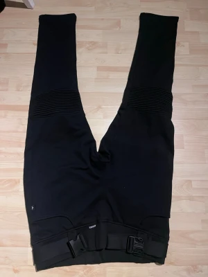 Svarta mc tights /byxor med spänne märket Krones  - Säljer ett par svarta MC-inspirerade byxor med ribbade detaljer över knäna och justerbart bälte i midjan. Byxorna har raka ben och två bakfickor. Perfekta för dig som gillar en sportig och tuff stil. Storlek 6XL. Inbyggt skydd i. Nya endast testade men klippt av lappen. Katt finns i hemmet 
