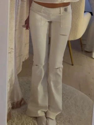Vita bootcut jeans från Juicy Couture - Säljer ett par vita bootcut jeans från Juicy Couture med låg midja och slitningar vid knäna. Jeansen har framfickor och en snygg metallknapp med logga. Perfekta för dig som vill ha en clean men edgy look.