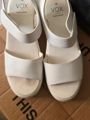 Vita platåsandaler från VOX - Säljer ett par vita platåsandaler från VOX med breda remmar och kardborrestängning. Sandalerna är veganska och har en chunky sula som ger extra höjd. Perfekta för dig som gillar minimalistisk och modern stil.