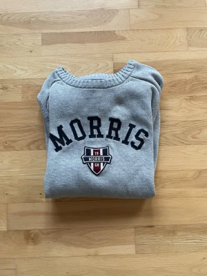 Morris - Grå stickad tröja från Morris med broderad logga och emblem på bröstet. Tröjan har rund hals, ribbade muddar och är tillverkad i mjukt stickat material. Perfekt för dig som gillar en klassisk look. 
