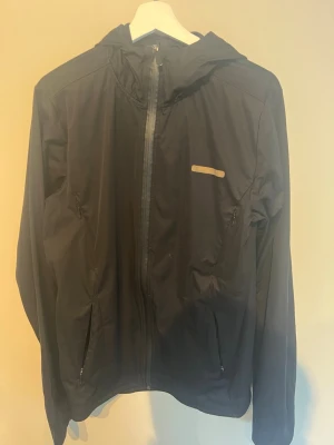 Svart regnjacka från A–Z med huva - Svart regnjacka från A–Z med hel dragkedja framtill, huva och två sidofickor med dragkedja. Jackan har en minimalistisk design med en liten reflekterande detalj på bröstet. Perfekt för regniga dagar och enkel att matcha med allt.