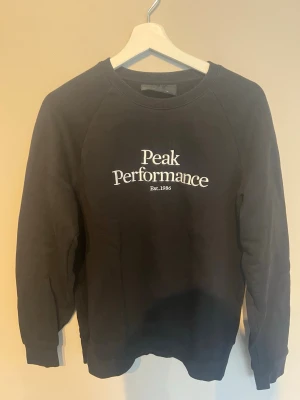 Svart Peak Performance sweatshirt - Cool svart sweatshirt från Peak Performance med vit broderad logotyp och text 'Est. 1986' på bröstet. Tröjan har rund halsringning och ribbade muddar vid ärmslut och nederkant. Perfekt för en avslappnad och sportig look.