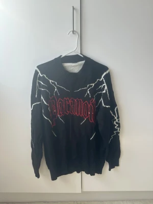 Svart stickad tröja med blixtar - Cool svart stickad tröja med vita blixtmönster och röd text 'Paranoid' på bröstet. Tröjan har rund hals och långa ärmar, perfekt för dig som gillar edgy streetstyle. Materialet är mjukt och bekvämt, passar till jeans eller cargo pants.