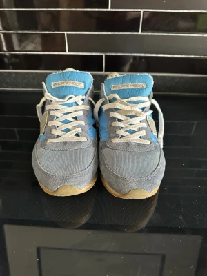 Philippe Model blå/grå sneakers - Snygga sneakers från Philippe Model i blått och grått med vita skosnören. Ovandelen är i mocka och textil, med en beige sula och logga på sidan. Perfekta för dig som gillar en sportig och avslappnad stil.
