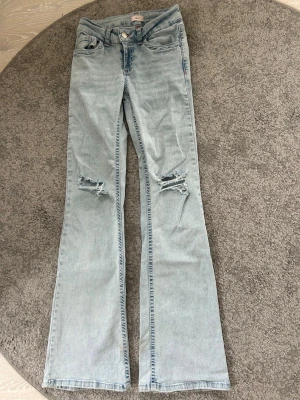 Ljusblå bootcut jeans med slitningar - Säljer ett par ljusblå bootcut jeans med slitna detaljer över knäna och broderade bakfickor med rosa zigzag. Jeansen har klassisk femficksmodell och är tillverkade i denim med en skön vintagekänsla. Perfekta för en avslappnad och trendig look. Något slitna där nere vid hälen, men inget man tänker på (se sista bilden)