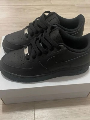 Svarta Nike Air Force 1 sneakers - Klassiska helsvarta Nike Air Force 1 sneakers i lågt utförande. Skorna har en stilren design med perforerad tå, svarta skosnören och diskret Nike Swoosh på sidan. Tillverkade i läder med gummisula och metall-detalj på snörningen.