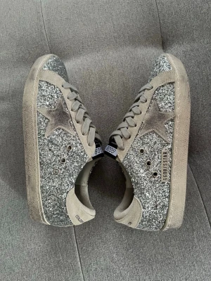 Golden Goose - Säljer ett par riktigt snygga sneakers från Golden Goose med silverglittrande paneler, beige mockadetaljer och den ikoniska stjärnan på sidan. Skorna har beige snörning och svart sula undertill. Perfekta för dig som vill sticka ut med stil!