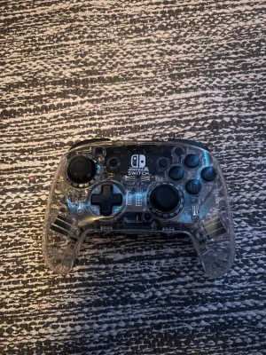 Nintendo Switch Pro Controller genomskinlig - Säljer en genomskinlig Nintendo Switch Pro Controller med svart detalj på baksidan. Handkontrollen har klassisk layout med två styrspakar, styrkors och alla vanliga knappar. Tillverkad i plast med transparent design som visar insidan. Perfekt för gaming på Switch!