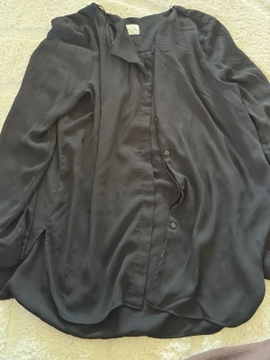 Svart silkeslen skjorta från Vila - Snygg svart skjorta från Vila i ett silkeslent material. Skjortan har klassisk krage, knappar framtill och lång ärm. Perfekt för dig som gillar en enkel men stilren look.