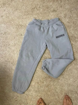 Gråa mjukisbyxor från Hoodrich - Säljer ett par gråa mjukisbyxor från Hoodrich med resår i midjan och vid bensluten. Byxorna har en relaxed fit och Hoodrich-loggan tryckt på ena benet. Perfekta för chill dagar och streetwear-stil.