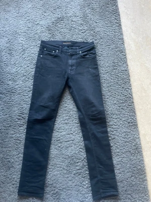 Svarta slim jeans från Nudie Jeans - Säljer ett par svarta slim fit jeans från Cheap Monday med klassisk femficksdesign och raka ben. Jeansen har metallknappar och är tillverkade i stretchigt material som ger en skön passform. Perfekta för dig som gillar en enkel och stilren look.