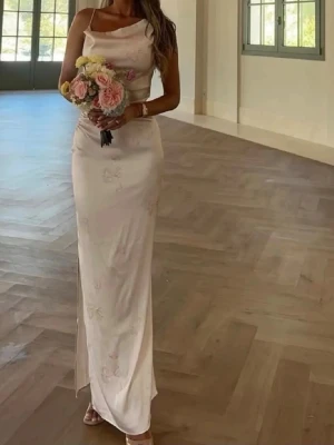 Ljusrosa långklänning med drapering - Elegant ljusrosa långklänning med smal passform och draperad axel. Klänningen har ett subtilt blommönster och är ärmlös med en axelbandslös design på ena sidan. Tillverkad i ett mjukt, glansigt material som ger en lyxig känsla.