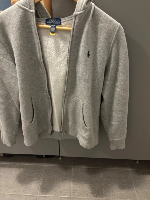 Grå hoodie från Polo Ralph Lauren - Snygg grå hoodie från Polo Ralph Lauren med dragkedja och två fickor framtill. Klassisk huva och diskret logga broderad på bröstet. Mjuk bomullsblandning som är riktigt skön att ha på sig. Perfekt för chill dagar eller när du vill ha något bekvämt.