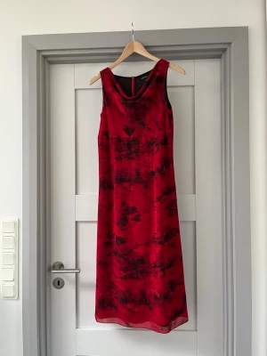 Röd ärmlös långklänning med mönster - Snygg röd långklänning med svart abstrakt mönster. Klänningen är ärmlös och har en rundad halsringning. Materialet ser ut att vara lätt och följsamt, perfekt för sommarens alla tillfällen. Ingen synlig logga eller märke.