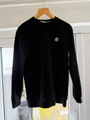 Svart sweatshirt från Moncler - Snygg svart sweatshirt från Moncler med rund halsringning och diskret Moncler-logga på bröstet. Tröjan har långa ärmar, ribbade muddar och är tillverkad i mjukt bomullsmaterial. Perfekt för en clean och stilren look.