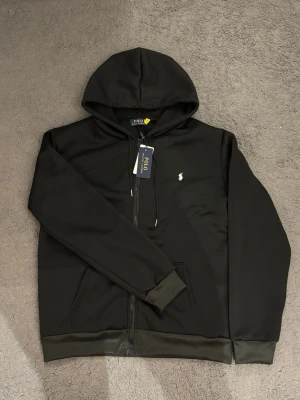 Ralph Lauren zip - Snygg svart hoodie från Polo Ralph Lauren med dragkedja framtill och klassisk huva. Ikoniska vita loggan broderad på bröstet. Tillverkad i mjukt material och har fickor på sidorna. Perfekt för en avslappnad och stilren look.
