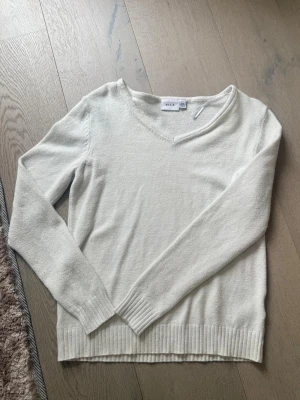 Vit V-ringad tröja från VILA - Klassisk vit tröja från VILA med V-ringning och långa ärmar. Tröjan har ribbade muddar vid ärmslut och nederkant, vilket ger en snygg och enkel look. Perfekt att matcha med jeans eller kjol för en clean stil.
