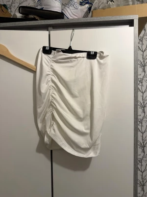 Vit kortkjol med rynkdetalj - Säljer en vit kortkjol med snygg rynkning på sidan som ger en cool draperad effekt. Kjolen har en enkel och stilren look, perfekt att matcha med olika toppar. Materialet känns mjukt och stretchigt.
