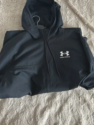 Svart windbreaker från Under Armour - Snygg svart hoodie från Under Armour med dragkedja och stor vit logga på bröstet. Tillverkad i ett mjukt och bekvämt material, perfekt för chill eller träning. Huva med dragsko för extra stil och funktion.