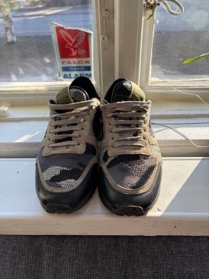 Valentino camo sneakers i grå och svart - Snygga sneakers från Valentino med camomönster i  svart och grått. Skorna har mockadetaljer, meshpaneler och en svart sula. Klassisk låg modell med snörning och logga på plösen. Perfekta för dig som gillar streetwear och exklusiva märken. Insidan är lite sliten men inget som stör eller syns när man bär de. Storlek 40 men passar 41 