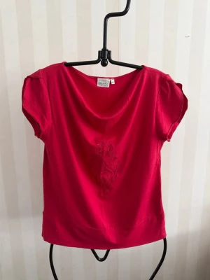 Röd blus med broderad detalj - Snygg röd blus från SOHO med korta ärmar och en broderad blomdetalj framtill. Blusen har en lös passform och mjukt fallande tyg som känns skönt mot huden. På etiketten storlek XL dock mer som en L.Perfekt för dig som vill ha en färgklick i garderoben!