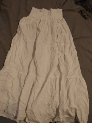 Vit långkjol med smock i midjan - Luftig vit långkjol med bred smock i midjan som ger en bekväm och flexibel passform. Kjolen har en lätt rynkad struktur och faller snyggt längs benen. Perfekt för varma dagar och enkel att matcha med olika toppar.