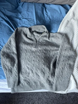 Grå kabelstickad tröja Polo Ralph Lauren - Säljer en grå kabelstickad tröja från Polo Ralph Lauren med halv dragkedja och hög krage. Tröjan har ribbade muddar vid ärmslut och nederkant, samt klassisk kabelstickad design. Perfekt för dig som gillar stilrena och tidlösa plagg. Änvänd max 5 gånger, säljer då den är lite för stor för mig