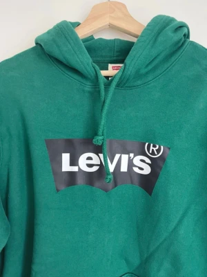 Grön Levi's hoodie med logga - Snygg grön hoodie från Levi's med stor vit logga på bröstet. Klassisk modell med huva och dragsko samt magficka framtill. Perfekt för en avslappnad och trendig stil. Tillverkad i mjukt material för extra komfort.