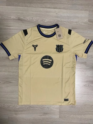 Fc Barcelona borta 25/26 - Snygg beige FC Barcelona matchtröja från Nike med svarta detaljer och tryck, inklusive Spotify-logga och klubbmärke på bröstet. Ribbade muddar med blå och svarta ränder, UNHCR-tryck på ryggen. Perfekt för fotboll eller träning.