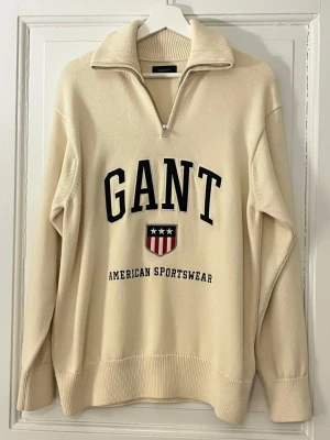 Beige stickad tröja från GANT - Snygg beige stickad tröja från GANT med halv dragkedja och hög krage. Tröjan har svart broderad logga och texten 'American Sportswear' framtill samt ett sköldmärke. Ribbstickade muddar vid ärmslut och nederkant. Perfekt för en sportig och avslappnad look. Aldrig använd, nypris är 1600kr. Kommer från fräscht djur och rökfritt hem