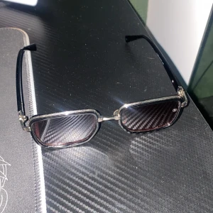 Svarta Balmain solglasögon  - Snygga solglasögon från Balmain med rektangulära svarta bågar och mörktonade linser. Metalliga detaljer vid gångjärnen och näskuddar för extra komfort. Perfekt accessoar för en trendig look.