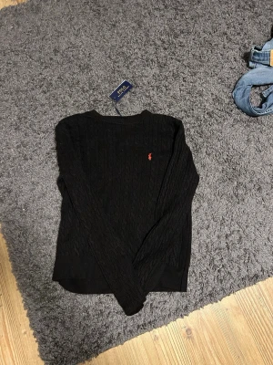Svart stickad tröja från Polo Ralph Lauren - Säljer en svart kabelstickad tröja från Polo Ralph Lauren med den klassiska röda loggan broderad på bröstet. Tröjan har rund halsringning och långa ärmar.  Hör av er vid funderingar pris kan diskuteras