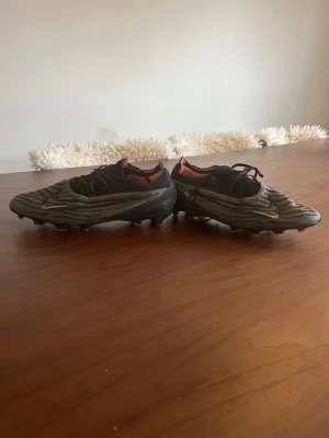 Nike phantom svart/grå fotbollsskor elite  - Säljer ett par Nike Mercurial fotbollsskor i svart och grått med unikt mönster och orange detaljer. Skorna har låg profil, snörning och dobbar för spel på gräs. Tillverkade i syntetmaterial för bra grepp och känsla på planen.
