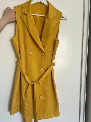 Gul ärmlös klänning med knappar - Snygg gul kortklänning med dubbelknäppning framtill och matchande bälte i midjan. Klänningen är ärmlös och har en klassisk krage, vilket ger en modern och stilren look. Perfekt för dig som vill sticka ut med färg och enkel design.