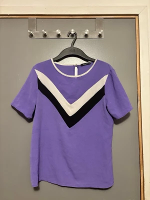 Lila topp med V-detalj köpt utomlands - Snygg lila topp från Busem (utomlands) med korta ärmar och rund halsringning. Framsidan har en tydlig V-formad detalj i svart och vitt som ger en unik vibe. Perfekt att matcha med jeans eller kjol för en cool look.