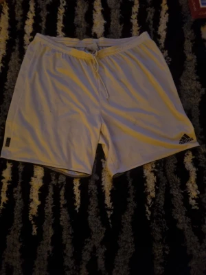 Vita Adidas shorts med snörning - Säljer ett par vita Adidas shorts med snörning i midjan och elastisk linning. Shortsen har en klassisk passform och är tillverkade i ett lätt och bekvämt syntetmaterial. Adidas-loggan syns på vänster ben. Perfekta för sport eller chill.