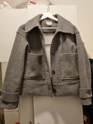 Grå ulljacka med stora fickor - Stilren grå ulljacka från Kappahl med bred krage, stora utanpåfickor och knäppning framtill. Jackan har en boxig passform och dekorativa knappar vid ärmsluten. Perfekt för dig som gillar minimalistisk och tidlös design. Tyvärr 2 söndriga knappar.