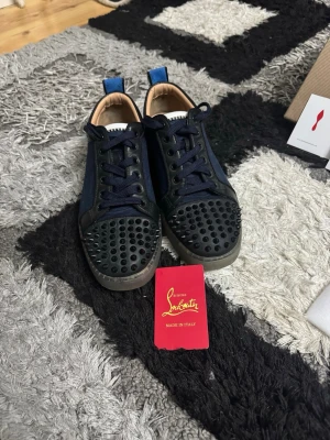 Christian Louboutin svarta och blå sneakers - Säljer ett par Christian Louboutin sneakers i svart och mörkblå med coola nitar på tån och klassisk röd sula. Skorna har snörning och är tillverkade i skinn och textil. Kommer med originalkartong och dustbag. Perfekt för dig som vill sticka ut med lyxiga detaljer.