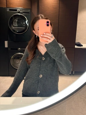 Mörkgrå kofta med knappar - Mysig mörkgrå kofta med stora, ljusa knappar framtill. Koftan har lång ärm och en enkel, rak passform. Perfekt att slänga på sig när det är lite kyligt. Materialet ser ut att vara stickat och mjukt.🎧🎉💖🍵😚