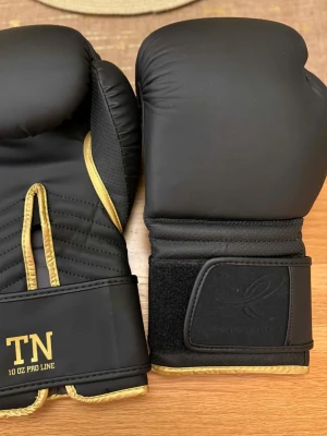 Svarta boxningshandskar TN 10 oz Pro Line - Snygga svarta boxningshandskar från TN med gulddetaljer och kardborrestängning. Tillverkade i syntetiskt läder och väger 10 oz, perfekta för träning eller sparring. Handskarna har ventilerande mesh och stilren design. Använd 2 gånger totalt.