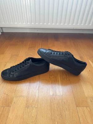 Svarta lädersneakers från Hugo Boss - Stilrena svarta sneakers i läder från Hugo Boss med diskret logga på sidan. Klassisk låg modell med svarta skosnören och svart sula. Perfekta för dig som gillar en clean och minimalistisk look. Skorna har en smal siluett och subtila detaljer.