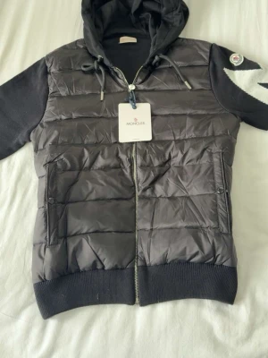 Svart pufferjacka med huva från Moncler - Svart pufferjacka från Moncler med huva och dragkedja framtill. Jackan har quiltad front i glansigt material, stickade ärmar och rygg samt två fickor med knappar. Vit detalj på ena ärmen och Moncler-logga på axeln. Perfekt för kalla dagar.