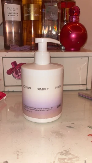 Lyko Simply Suede lotion - En stilren pumpflaska med LYKO Simply Suede lotion. Flaskan är vit med lila ombre-effekt och har svart text. Lotionen har en doft av bergamott, varm trä och vit mysk. Perfekt för att återfukta huden och ge en fräsch känsla.