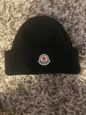Monclermössa - Säljer en svart ribbstickad mössa från Moncler med uppvikt kant och klassisk Moncler-logga framtill. Mössan är i tjockt, mjukt material och har en enkel, stilren design som passar perfekt till kalla dagar.