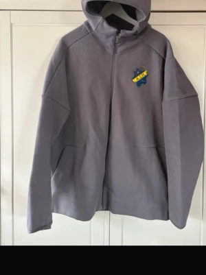 Grå AIK hoodie från Adidas - Snygg grå hoodie från Adidas med AIK-logga på bröstet och Adidas-logga på ärmen. Tröjan har dragkedja framtill, huva och är tillverkad i ett mjukt material som känns skönt mot huden. Perfekt för dig som vill visa ditt AIK-stöd med stil.