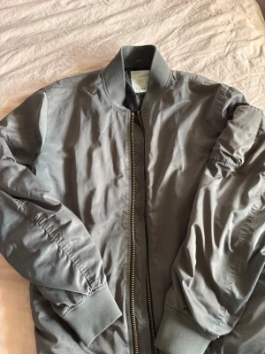 Grå bomberjacka från J.Lindeberg - Snygg grå bomberjacka från J.Lindeberg med klassisk ribbad krage, muddar och dragkedja framtill. Jackan har en minimalistisk look och är tillverkad i ett glansigt material som ger en modern vibe. Perfekt för dig som vill ha en stilren och trendig jacka.